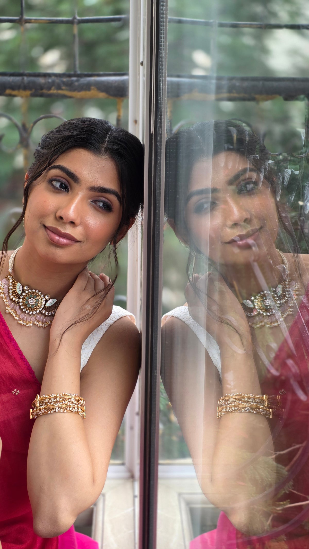 A woman in a pink saree leans on a window, her polki choker and bangles mirrored in the glass